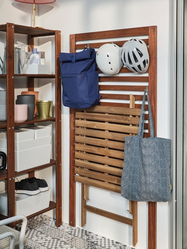 Una mesa de madera ASKHOLMEN plegada y colgada de un panel mural para terraza ÄPPLARÖ, bajo dos cascos y una mochila. Junto al panel hay un estante de madera con maceteros vacíos y cajas organizadoras. 