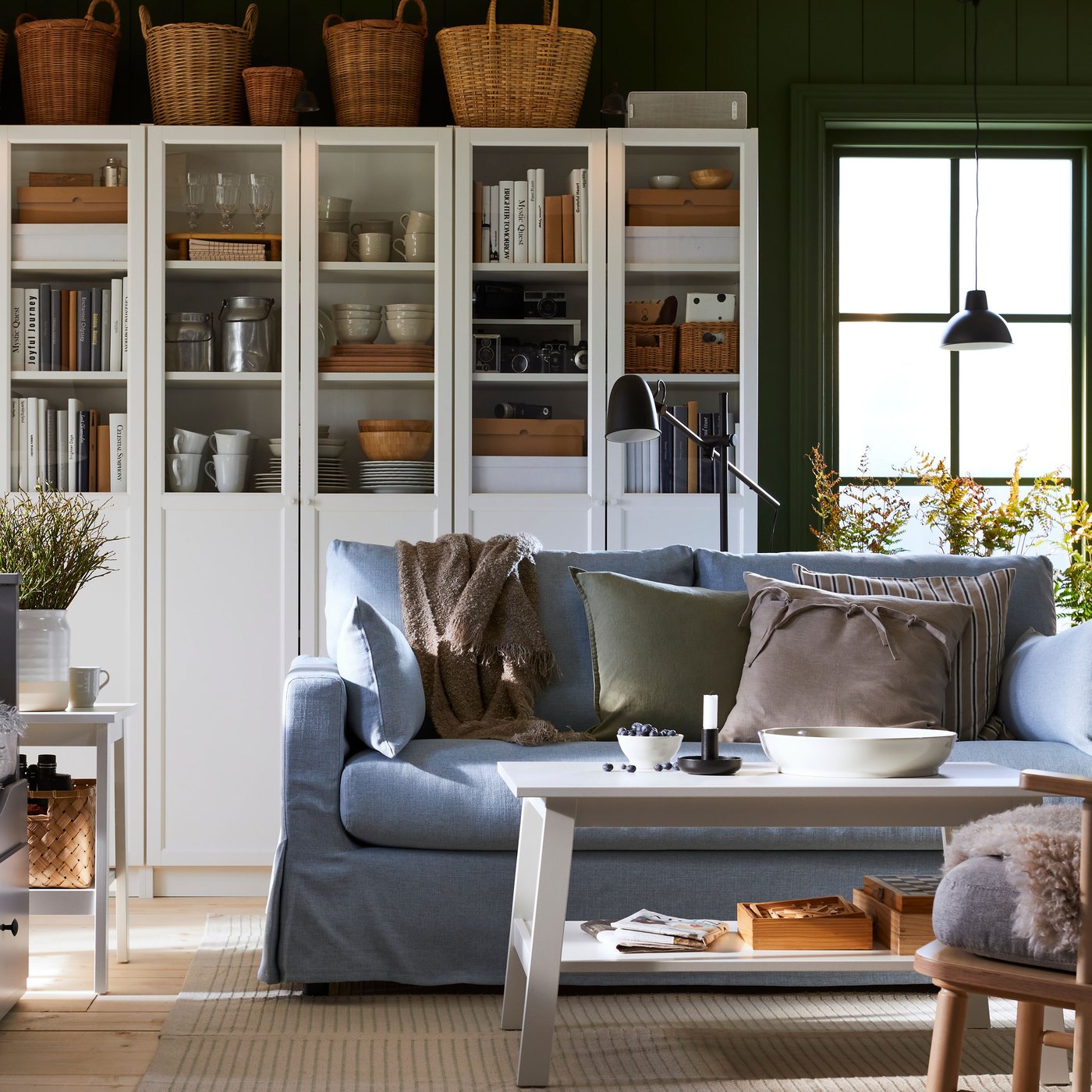 Librerías BILLY blancas con paneles/puertas de vidrio llenas de libros, tazas y platos, detrás de un sofá azul y una mesa de centro blanca.