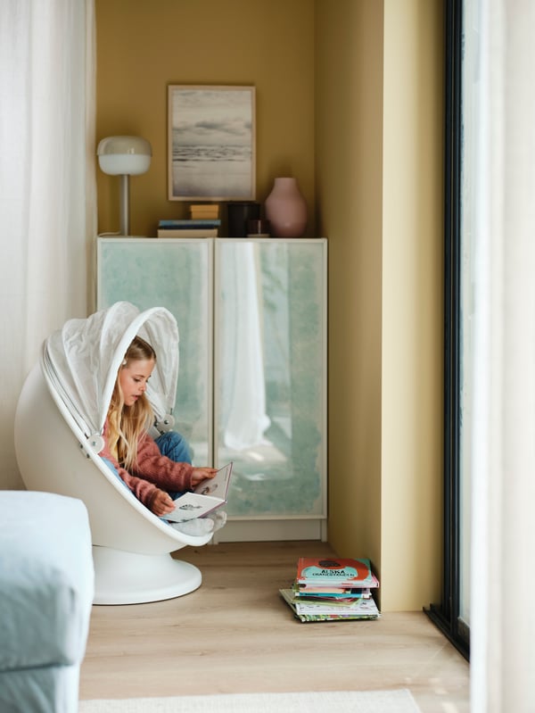 A child is sitting and reading inside an IKEA PS LÖMSK swivel armchair next to a bookcase combination with glass doors.