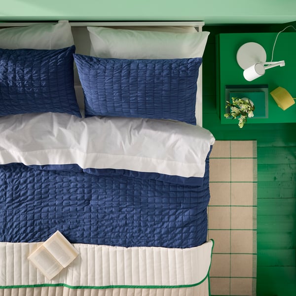 A work lamp with wireless charging sits on a green bedside table beside a bed with a dark blue RÄVSTARR comforter set.