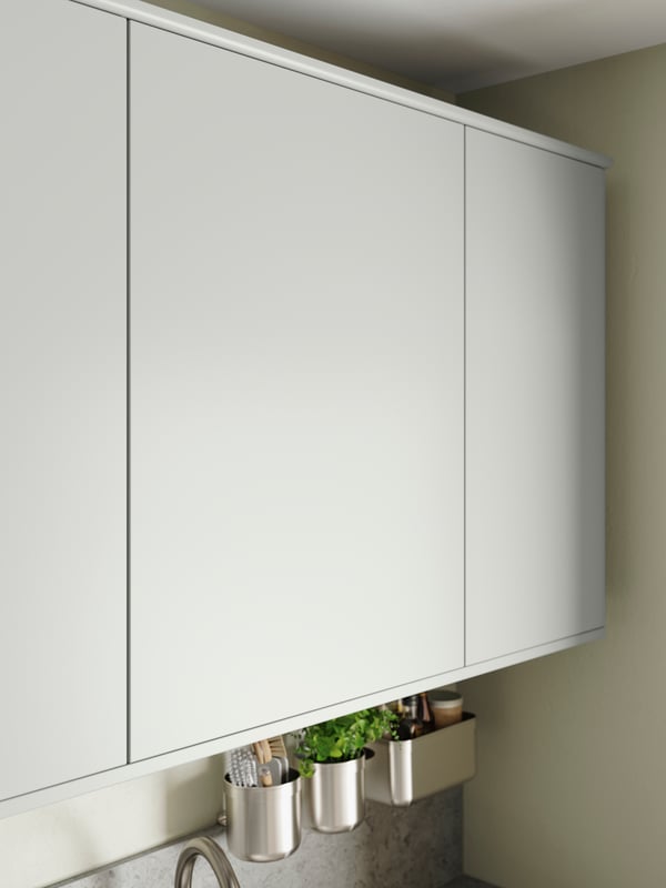 Three wall-mounted cabinets with smooth, light grey HAVSTORP fronts have a KUNGFORS rail with hooks in stainless steel below.