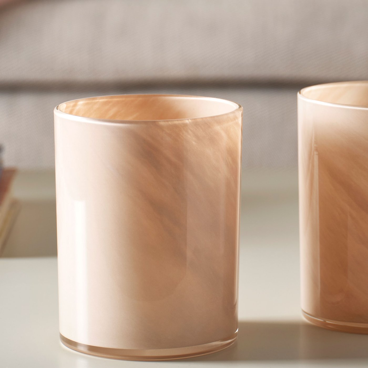 Närbild på två beiga VINDSTILLA ljuslyktor stående på bitt bord framför en meige soffa. 