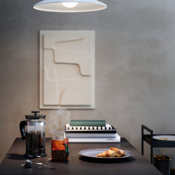 A dining table with an UPPHETTA coffee/tea maker, a mug, a croissant, and a pile of books, underneath an abstract art piece.