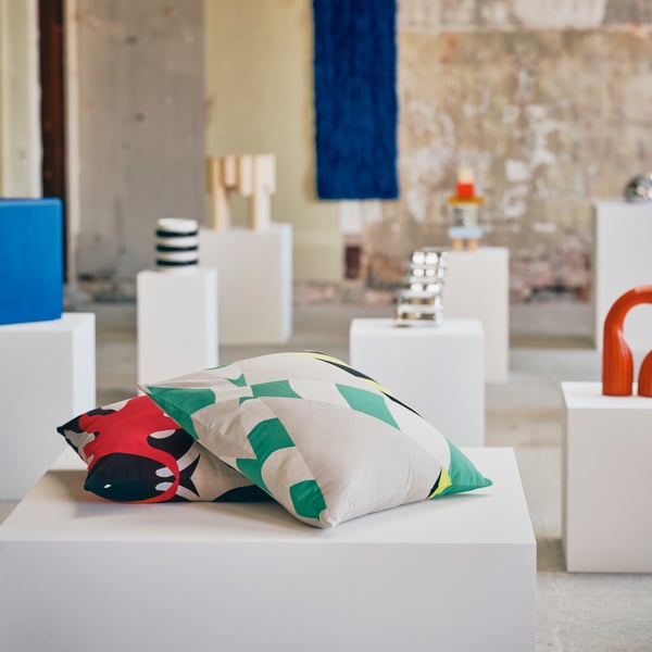 A rough gallery style room with IKEA art objects on white pedestals. In the foreground are two colourful pillows.