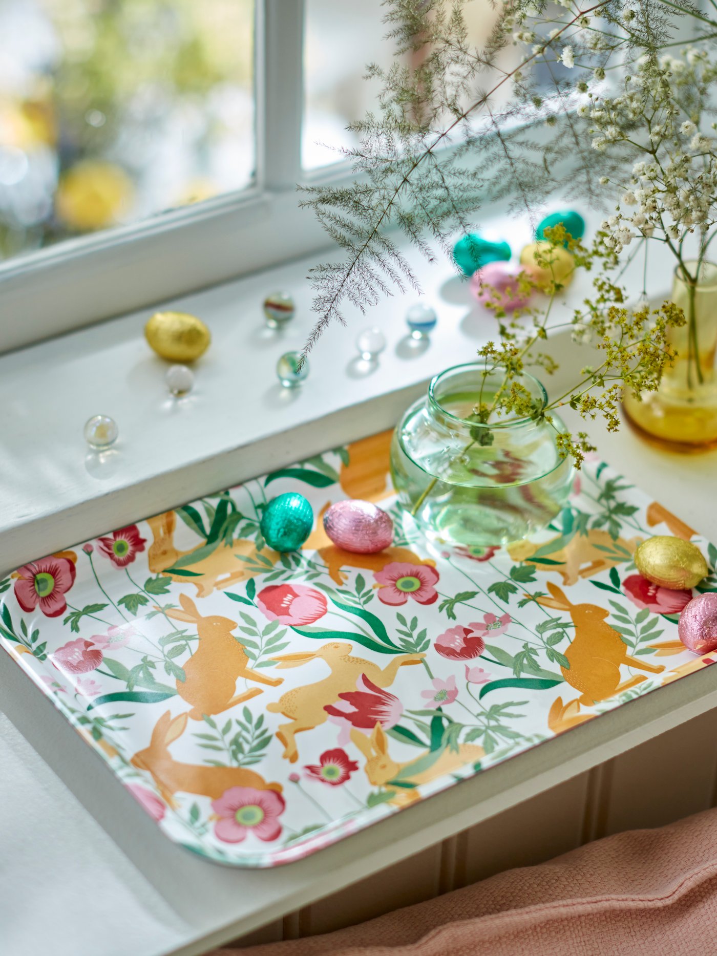 A floral-patterned TJÄRLEK tray by a window, carrying VÅRKÄNSLA chocolate eggs and a vase with SMYCKA artificial sprays.