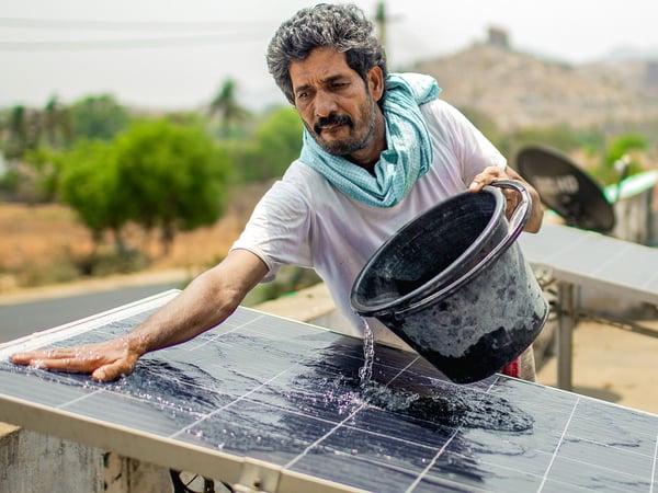 A person is cleaning a solar panel. The individual is using their right hand to spread water or a cleaning solution over the surface of the panel. The background features a blurred natural landscape, emphasizing the maintenance of sustainable energy sources.