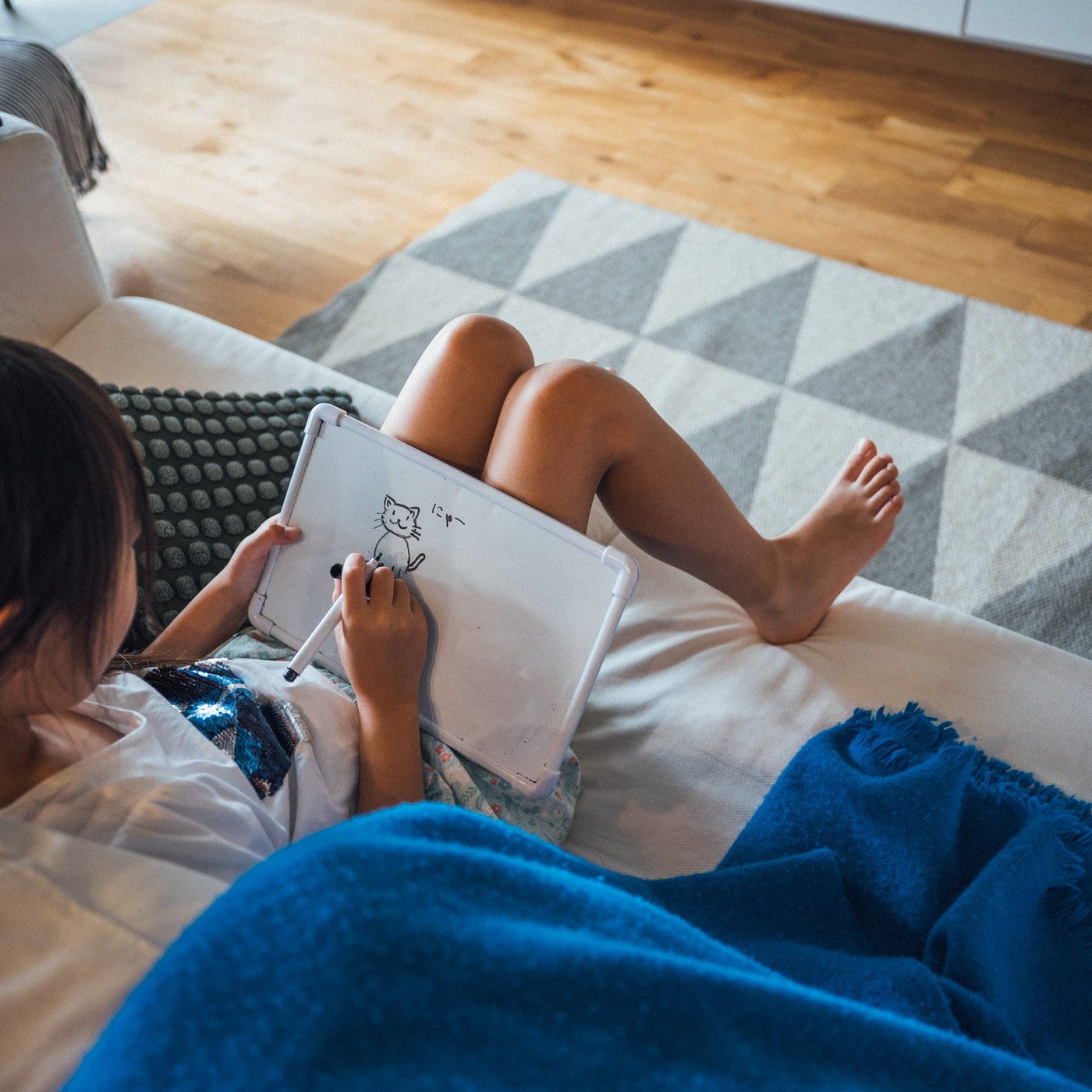 白いソファに座った子どもがひざのボードに絵を描き、足元には三角模様のラグと青いブランケットがあ
