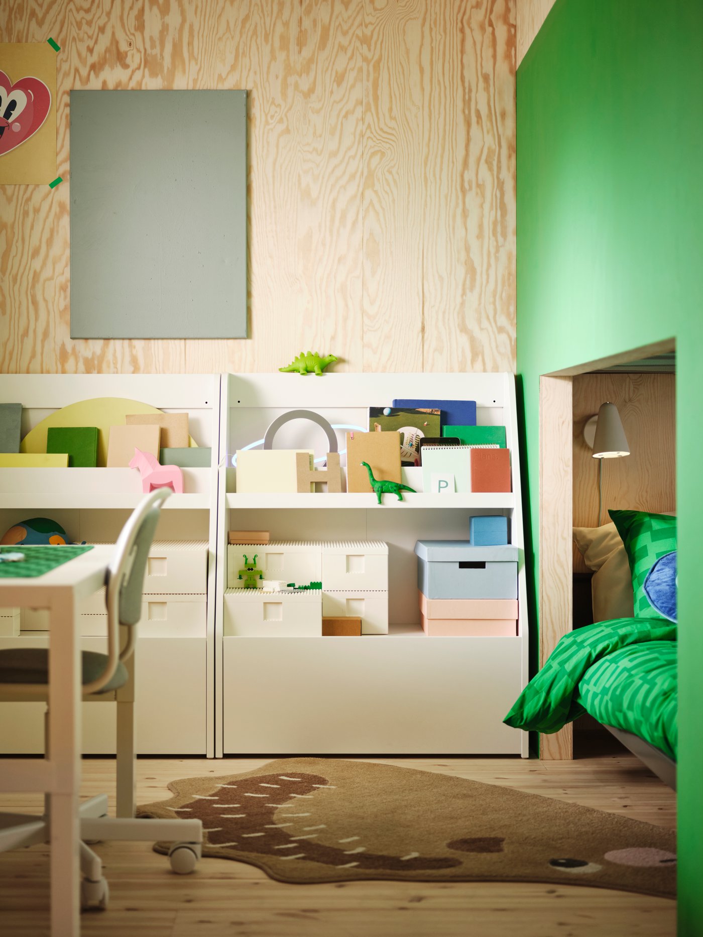 Contra una pared hay dos estantes de libros BERGIG blancos con espacio de almacenamiento, que contienen una variedad de cajas, libros y juguetes.
