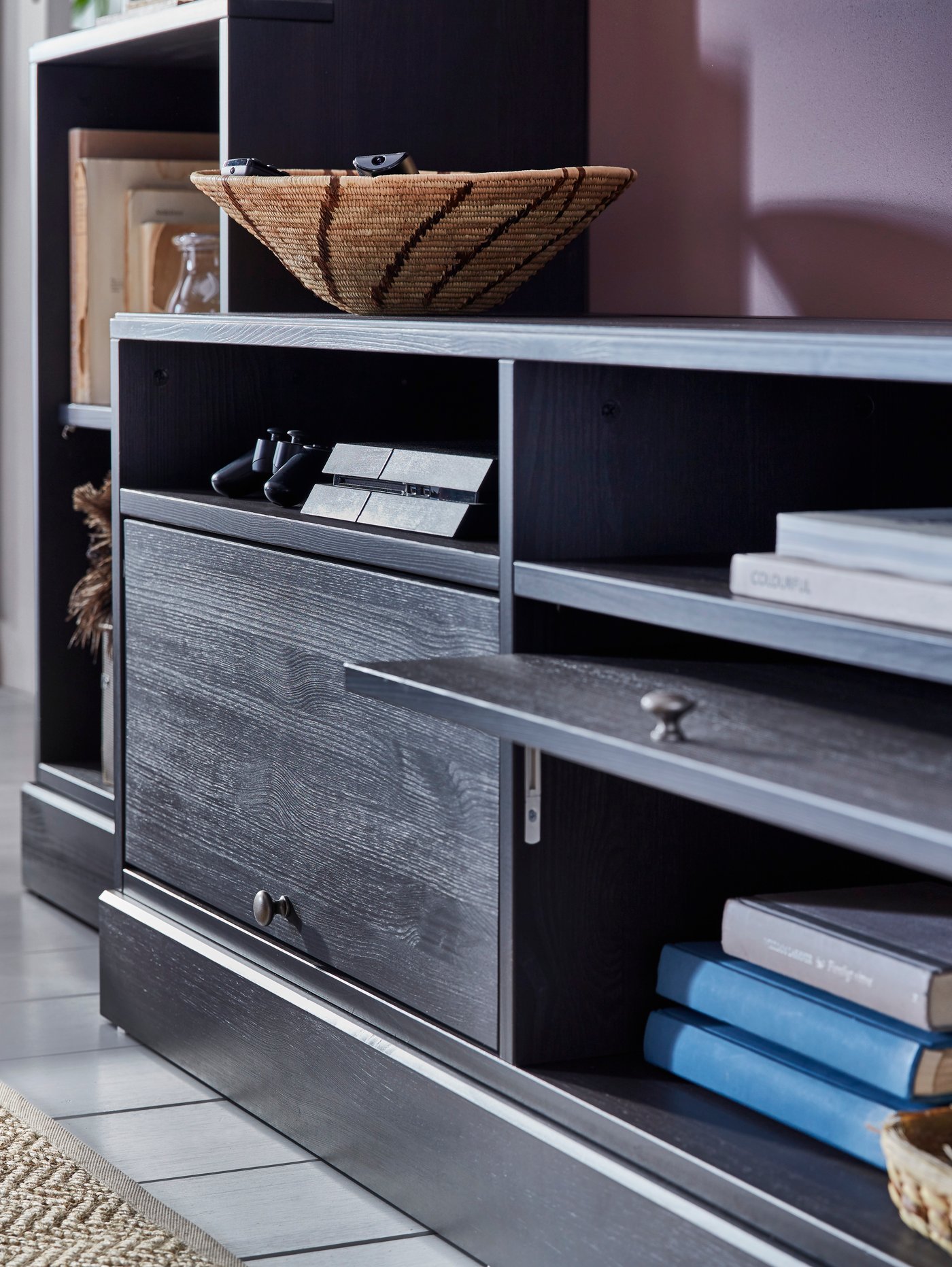 Detailansicht eines dunklen IKEA HAVASTA TV-Boards mit aufgeklappter Tür, offenen Fächern mit Büchern und Fernbedienungen sowie dekorativer Schale – modernes Wohnzimmermöbel vor rosafarbener Wand.