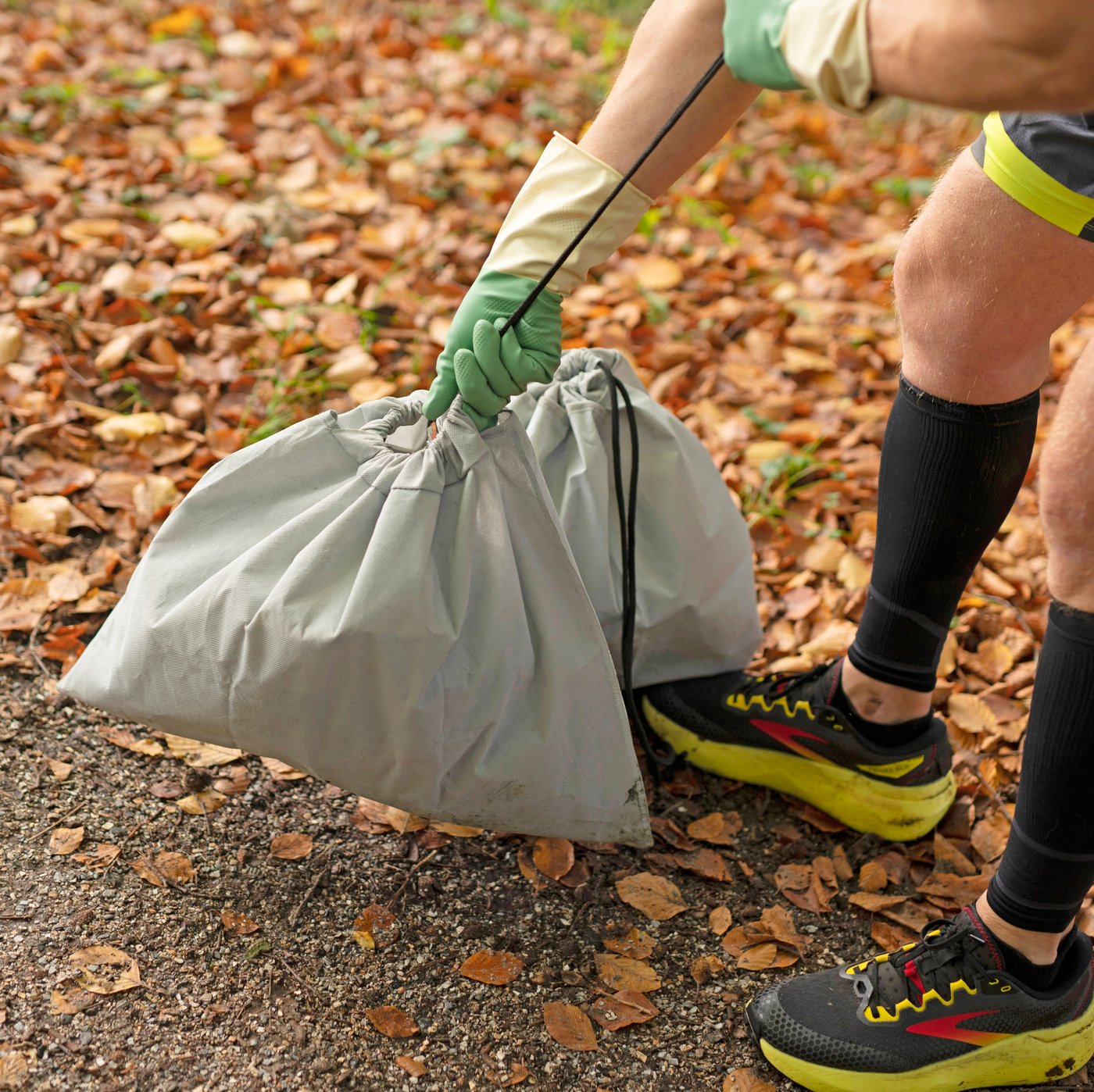 A man outdoors, wearing sports clothes and RINNIG cleaning gloves, ties up an AJÖSS waste sorting bag, filled with litter.
