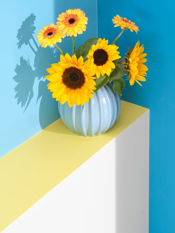 A light blue SKOGSTUNDRA vase filled with yellow sunflowers and gerberas.