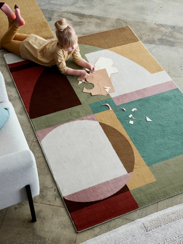 STENMÄTARE rug with a Bauhaus pattern in pastel tones lying by a sofa with a girl on the rug doing arts and crafts.