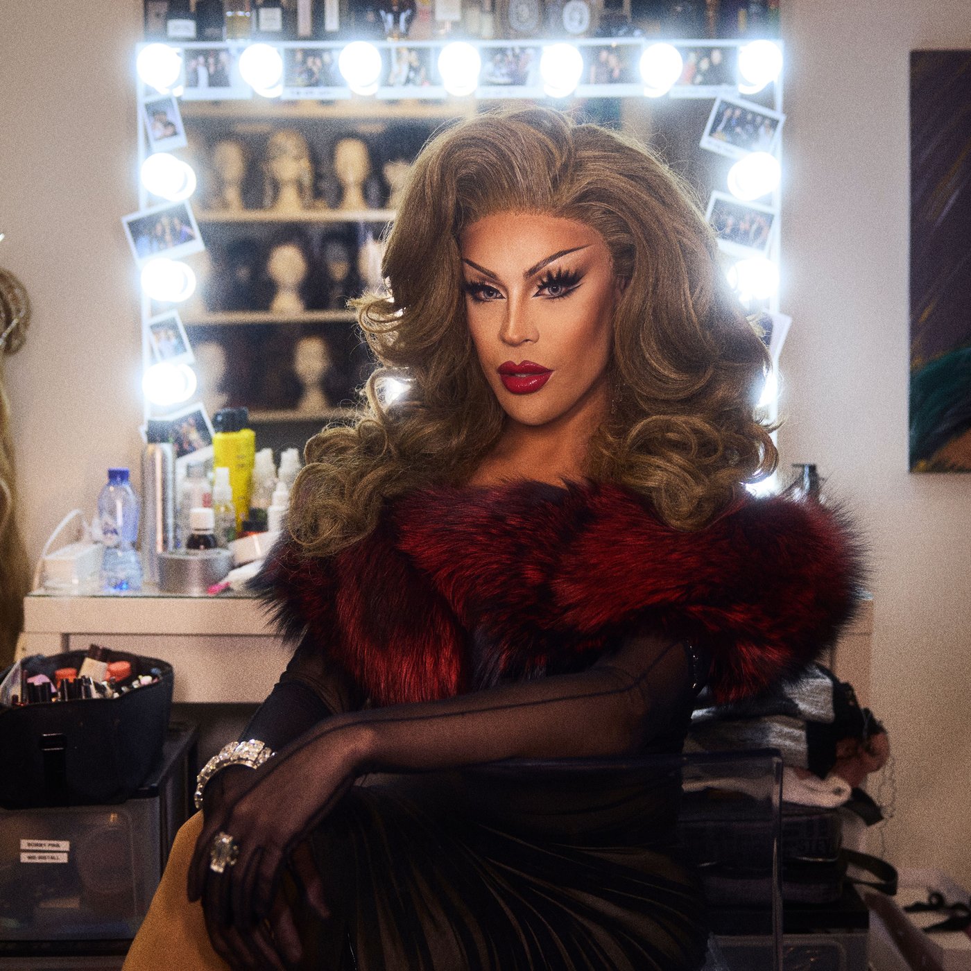Envy Peru sitting in full make up and drag in her dressing room, the vanity mirror behind casts a glamorous shine.
