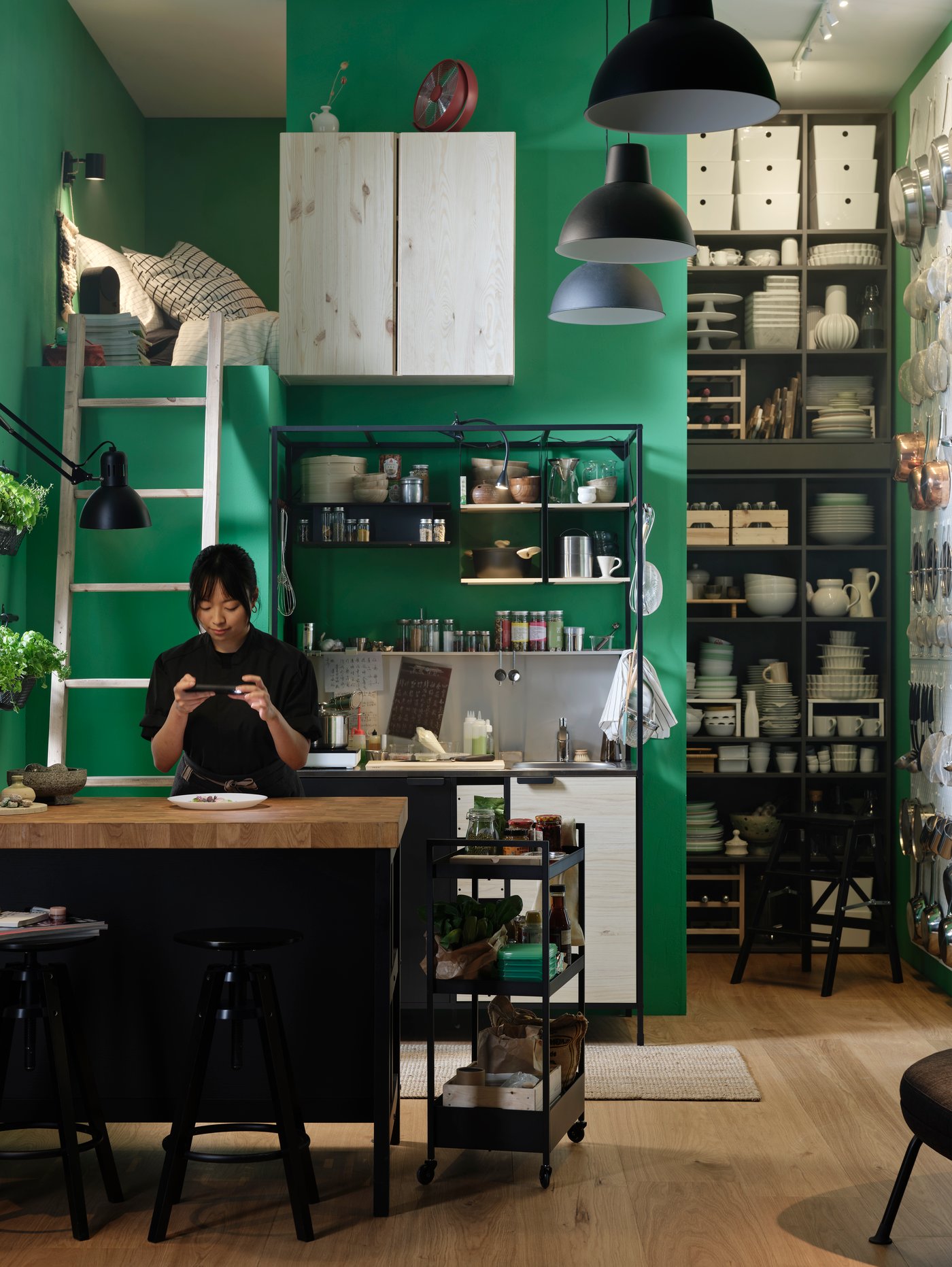 A person at a kitchen island photographs food on a plate. An ÄSPINGE kitchenette is placed by a green wall in the background.
