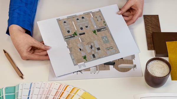 Projektantka wnętrz IKEA prezentująca kartki z wydrukiem rzutu z góry zaprojektowanych pomieszczeń biznesowych. Obok na blacie leży wzornik kolorów i próbki materiałów.