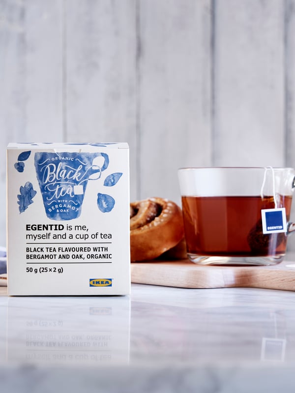 Cinnamon roll and tea in glass cup on small tray, in turn placed on a section of marble worktop; packet of tea on the side.