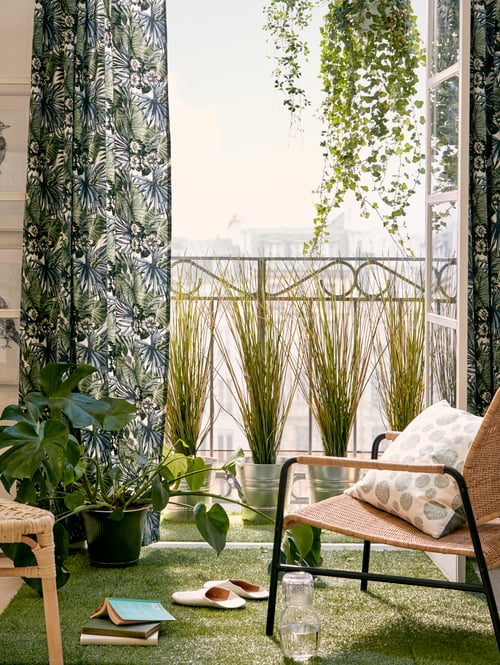 Living room featuring an armchair in front of an open window, with green curtains, plant pots, and a stool.