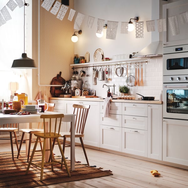 A white kitchen in traditional style. Several wooden dining chairs are around the table and bunting hangs from the ceiling.