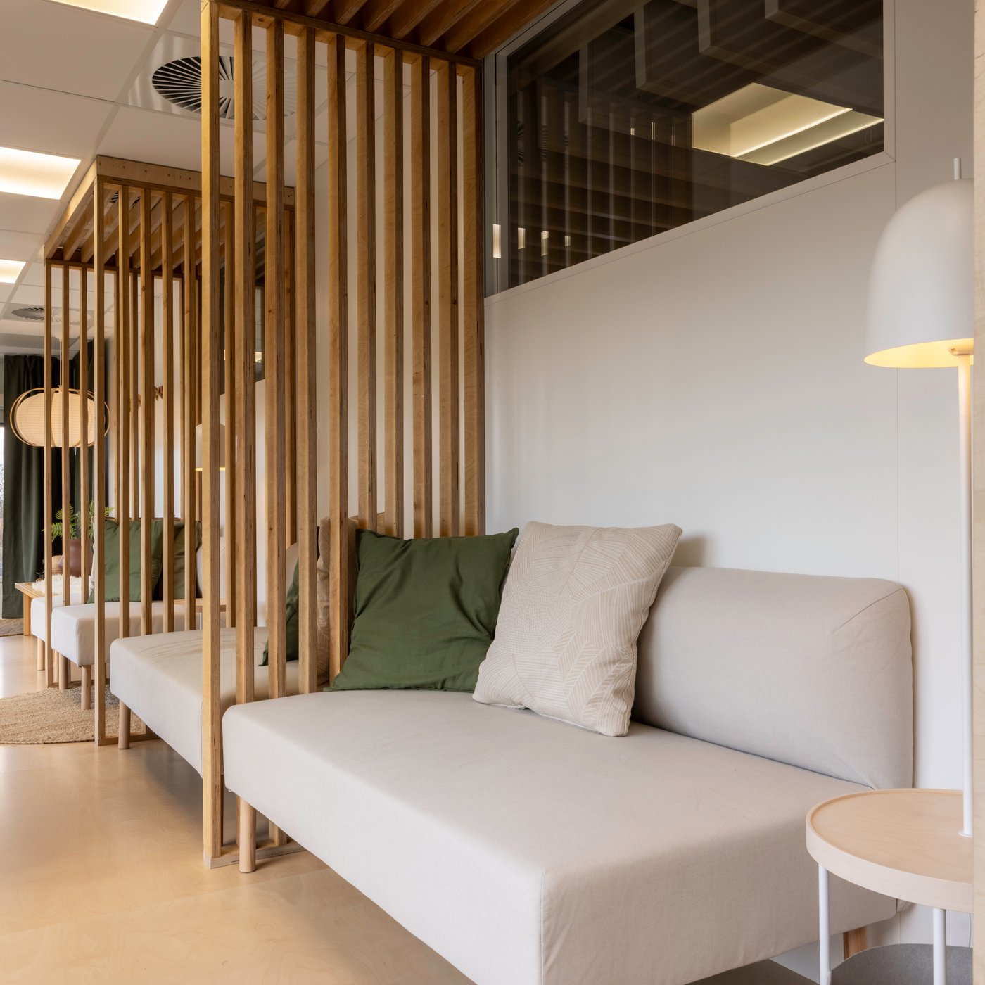 Espace lounge de bureau serein avec un canapé blanc, un coussin vert et des cloisons en lattes de bois qui créent un coin calme et semi-privé.