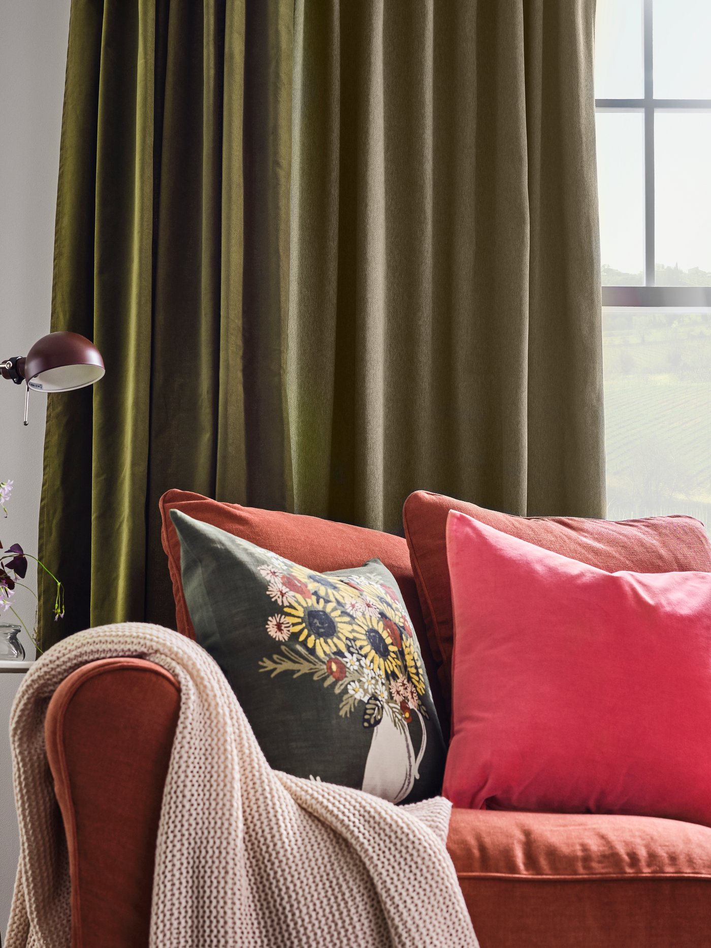 Light red sofa with cushions in different colours and a pale pink throw. Green curtains hang at the window behind.
