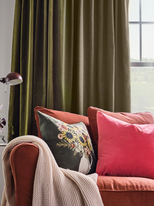Light red sofa with cushions in different colors and a pale pink throw. Green curtains hang at the window behind.