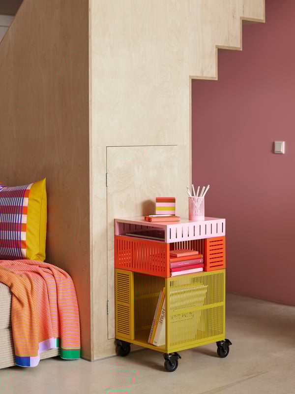 A colourful TESAMMANS storage unit on castors has books in it and stands behind a wooden staircase close to a pink wall.