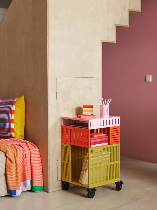 A colourful TESAMMANS storage unit on castors has books in it and stands behind a wooden staircase close to a pink wall.