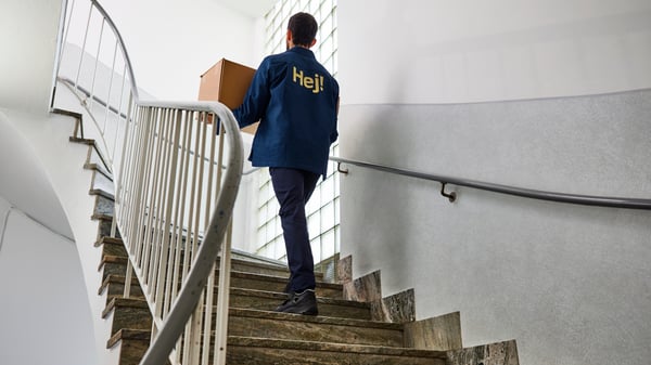 Person wearing an IKEA uniform carrying a box up a staircase in a bright building.