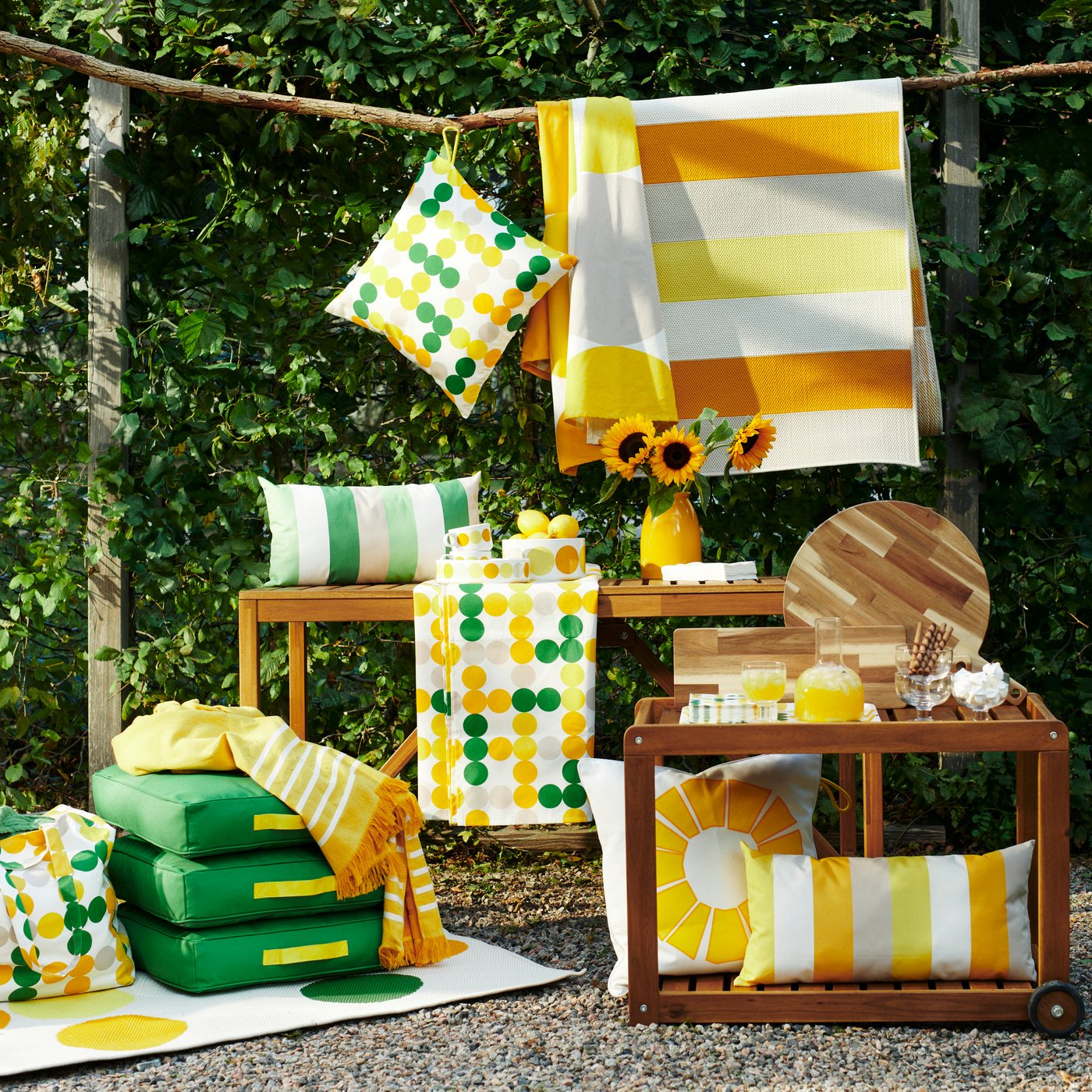 A collection of colourful BRÖGGAN outdoor textiles in a greens and yellowss displayed on two outdoor wood tables