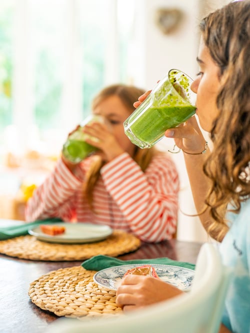 Two young girls sitting at a dining table drinking freshly made green smoothies from POKAL glasses.