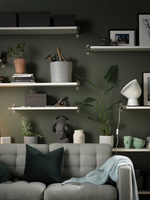 A sofa in a living room with six shelves on a grey wall. They hold boxes, photographs, lamps, plant pots, and other objects.
