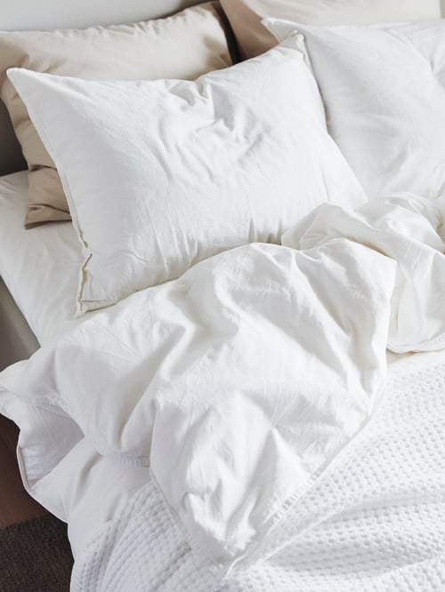 Bedroom with white duvet cover and two pillowcases, white bedspread, beige cushion covers, and bed frame with headboard.