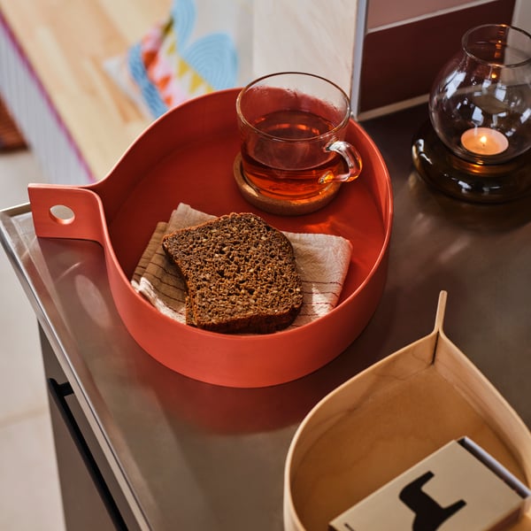Vista dall’alto di un vassoio OMMJÄNGE arancione con all’interno una bevanda in tazza trasparente e una fetta di plumcake appoggiata sopra un tovagliolo.
