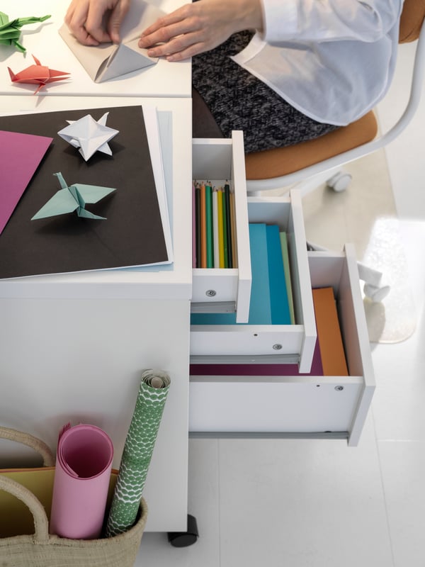 Una cajonera Micke con papeles de colores en el interior y al costado una niña haciendo origami con ellos.