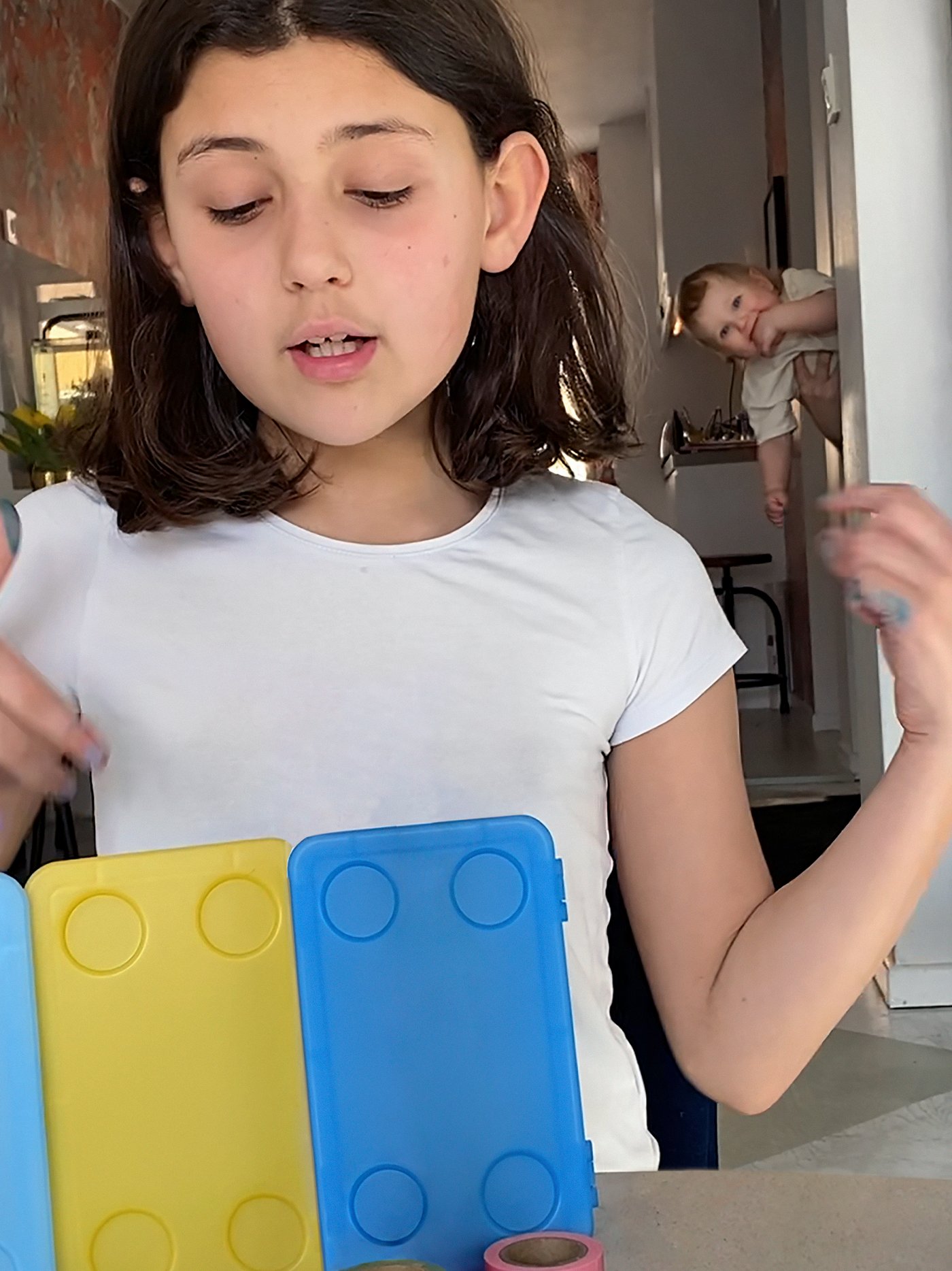 A girl looks down at three GLIS boxes. In the background there is a baby, held by an adult.