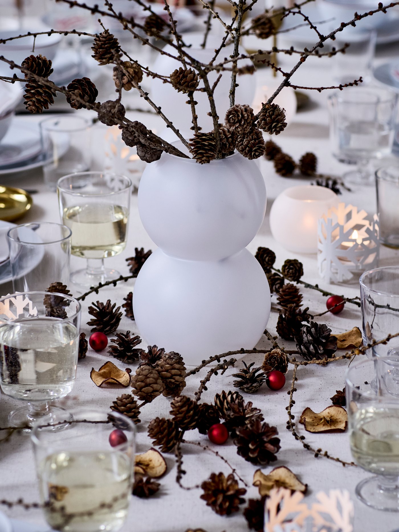 A white-themed dinner table is decorated for Christmas with a VINTERFINT vase with branches in it as a centrepiece. 