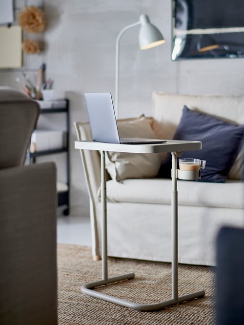 Un ordinateur portable sur un support pour ordinateur portable BJÖRKÅSEN beige, un tapis en jute, un fauteuil avec des coussins gris foncé et un lampadaire/lampe de lecture blanc.