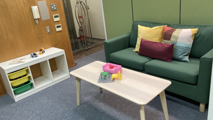 緑のソファとカラフルなクッション、ローテーブルとおもちゃ、収納付きシェルフがあるリビングルームのキッズスペース