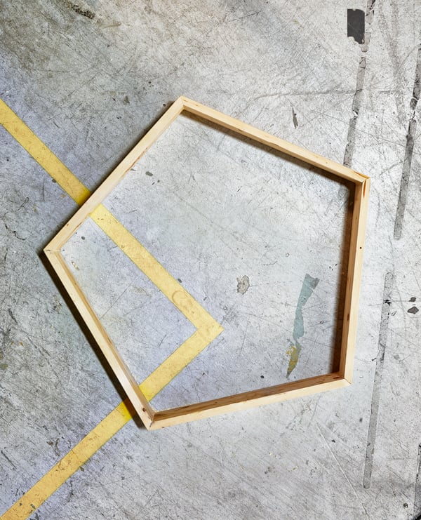 A wooden frame in the shape of a pentagon lying on a scuffed concrete floor.