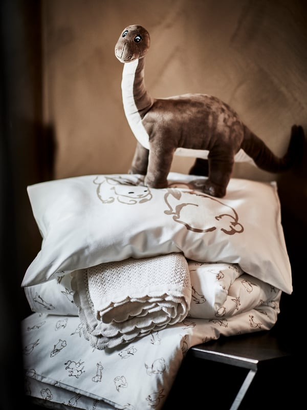 A JÄTTELIK soft toy shaped like a dinosaur stands on top of a pillow and duvet in RÖDHAKE covers and a GULSPARV blanket.