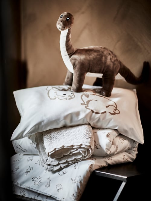 A JÄTTELIK soft toy shaped like a dinosaur stands on top of a pillow and duvet in RÖDHAKE covers and a GULSPARV blanket.