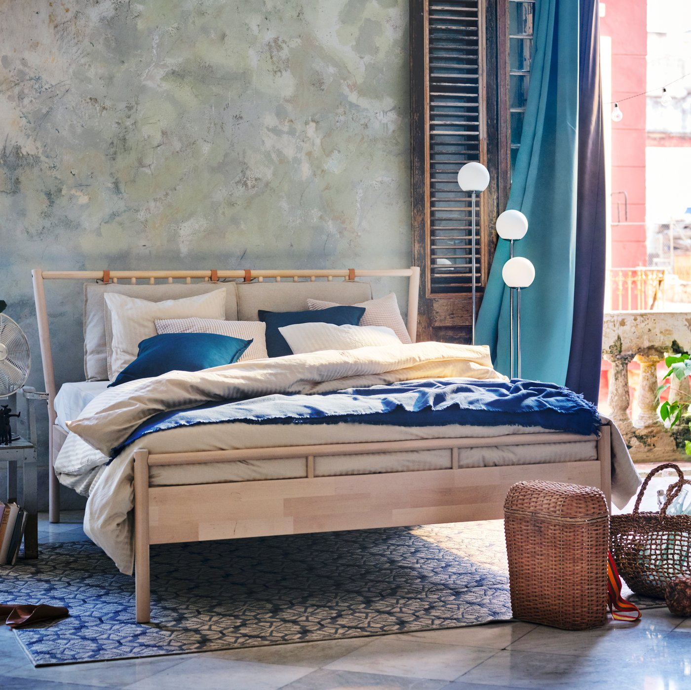 Un dormitori de tons blaus i grisos amb un llit doble de fusta i cortines opaques de color turquesa i morat que es mouen amb el vent.