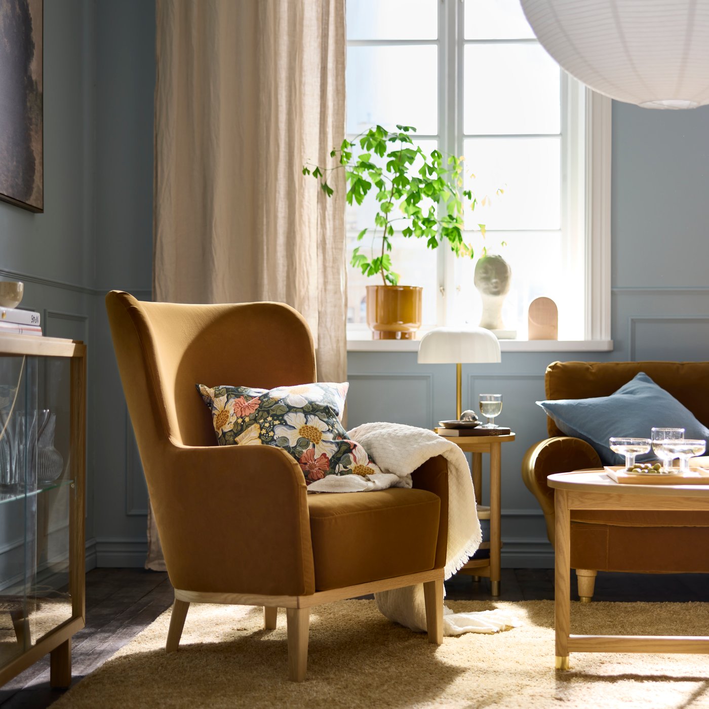 By a window in a sunlit living room is a dark-yellow-brown FAGERBACKEN armchair with a floral-pattern cushion in it.