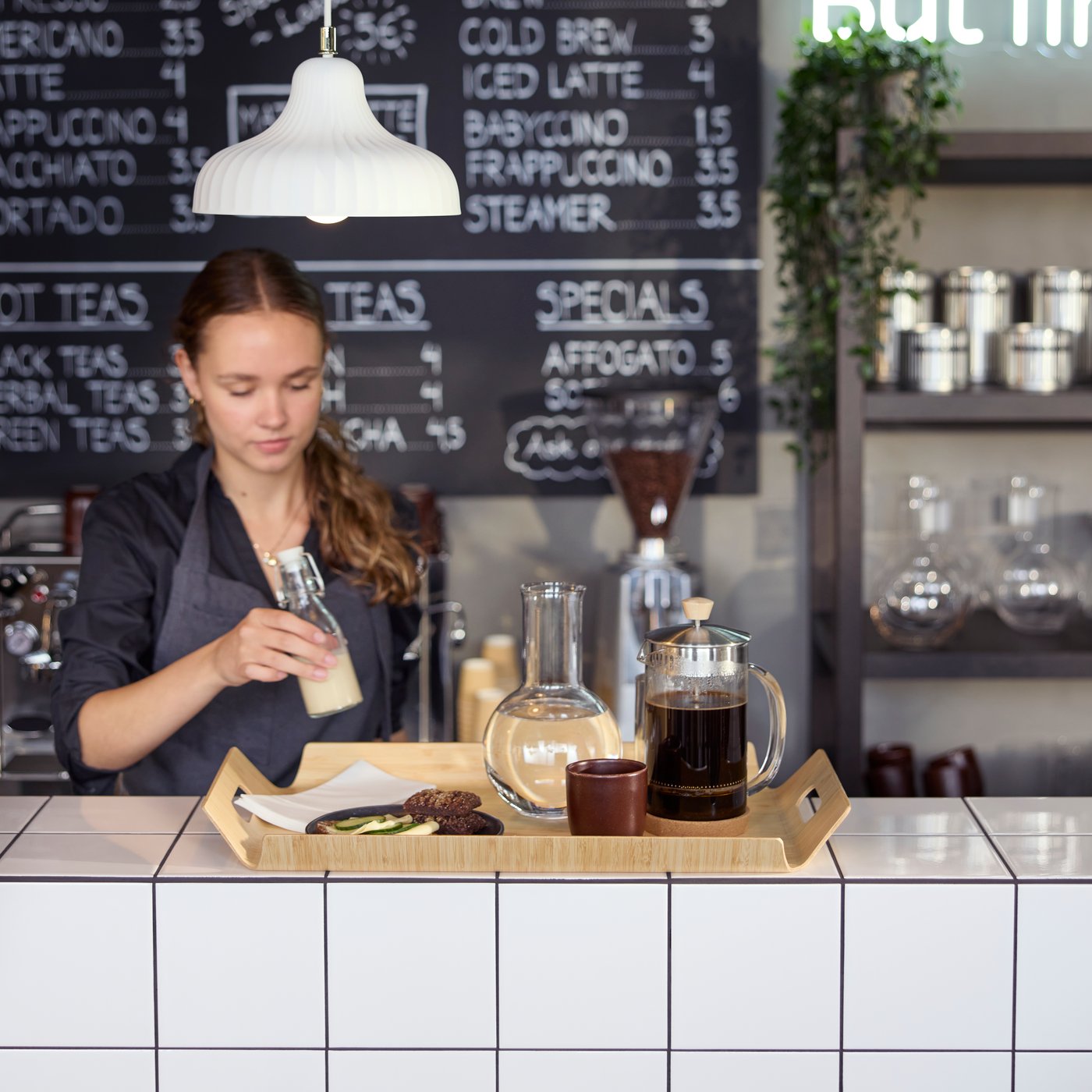 Café scene with a barista preparing a bamboo SILVERHAJMAL tray to serve drinks and food on a tiled café counter.