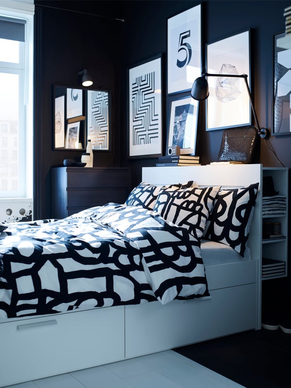 Bedroom with black walls, white wooden bed frame with drawers and shelves, black and white bed linens, and framed posters.