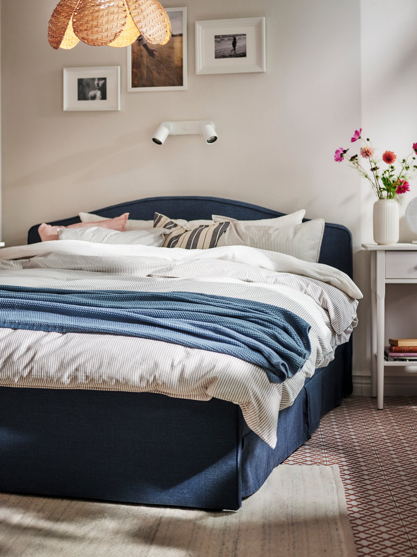 Niebieskoszary pled HUMLEMOTT leży na tapicerowanym łóżku RAMNEFJÄLL nakrytym pościelą SCHERSMIN i SOLFIBBLA. 