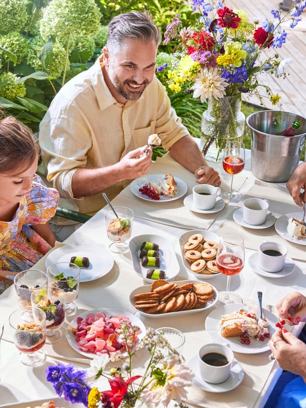 Eine Familie feiert ein schwedisches Mittsommerfest im Freien. Alle sitzen um einen Esstisch mit vielen Snacks, Kuchen und Kaffee.