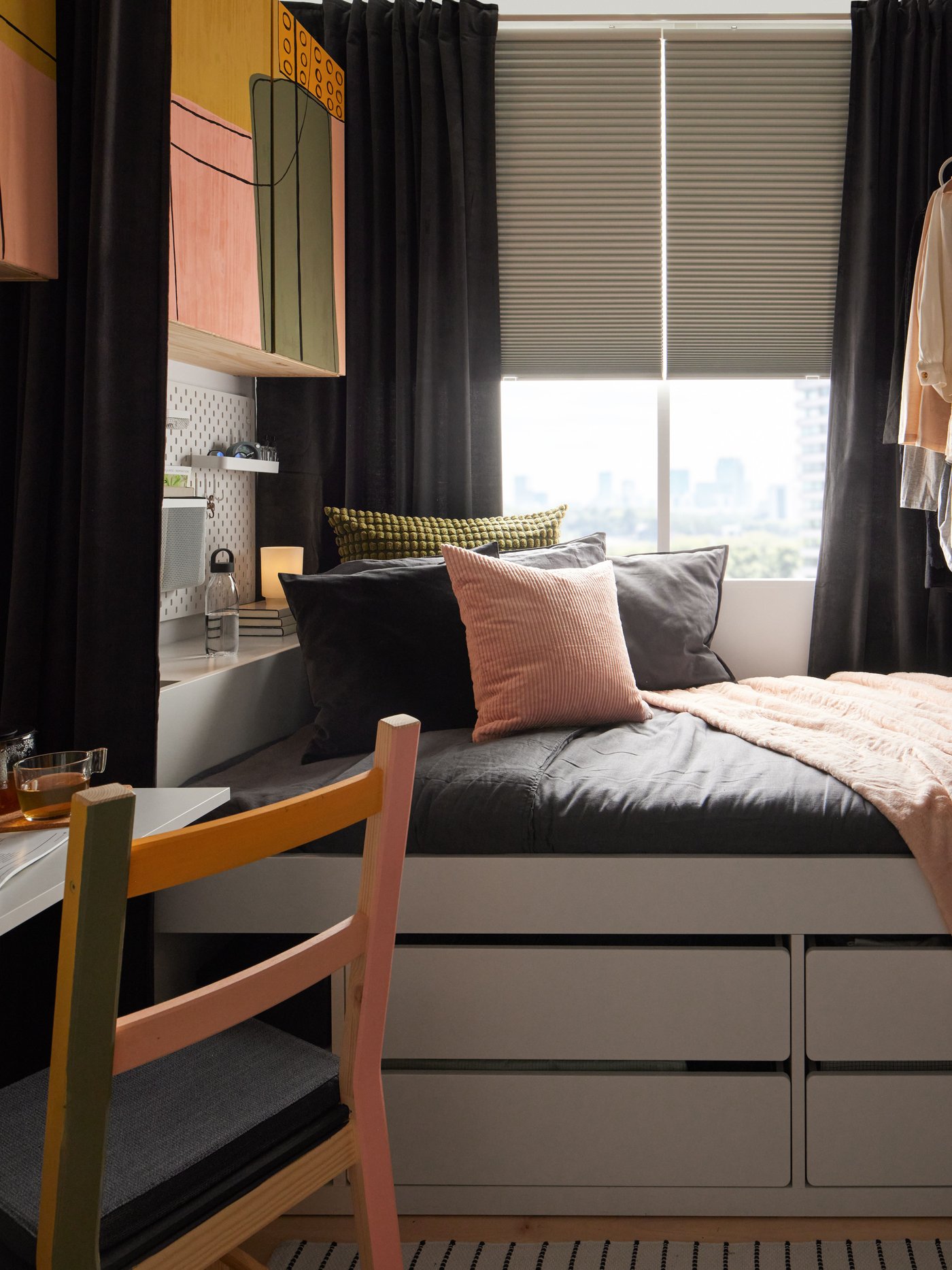 A view of a bed placed by a window half covered with blinds. A chair in the front and a cupboard over the bedhead are painted in yellow, pink and green.
