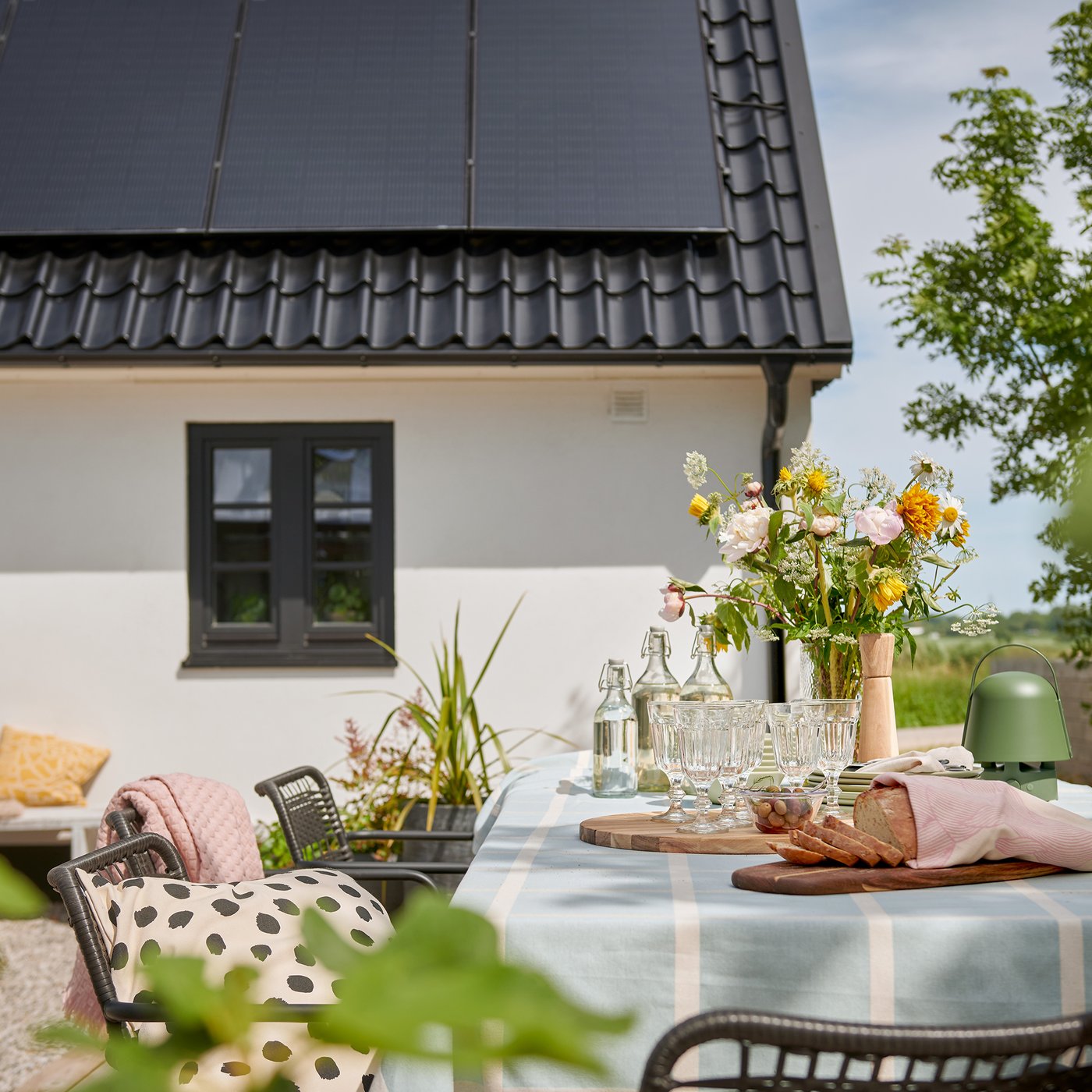 Eine sonnige Essbereich im Freien mit einem Tisch voller Blumen und Erfrischungen, vor dem Hintergrund eines Hauses mit Sonnenkollektoren.