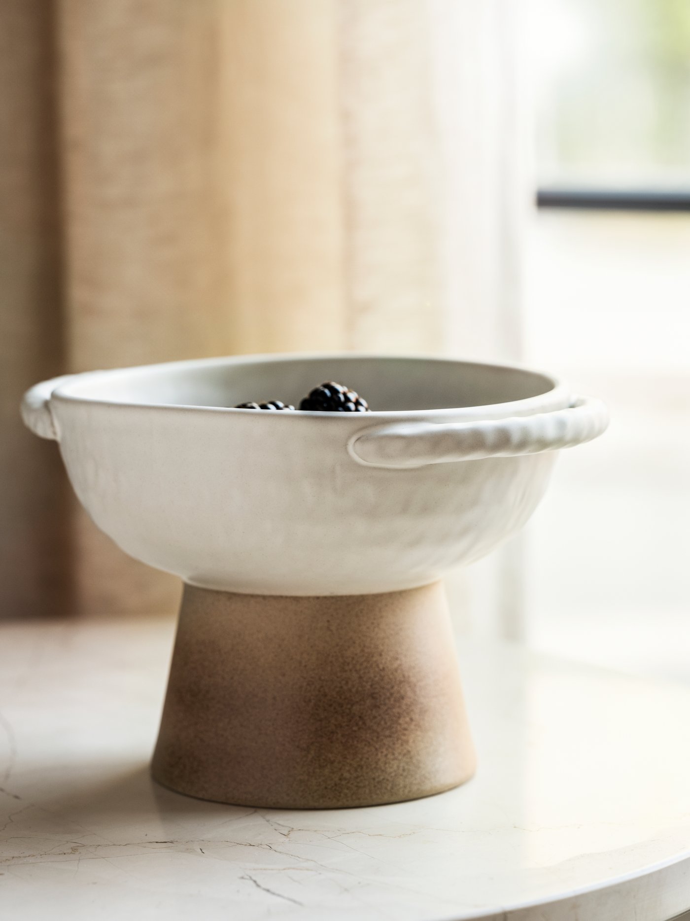 A handmade, tall ceramic bowl with two handles, placed on a marble surface in front of a window.